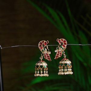 Może przedstawiać: Para złotych kolczyków z czerwonymi i zielonymi kamieniami szlachetnymi. Kolczyki mają kształt pawia i wiszącą ozdobę w kształcie dzwonka z białymi perełkami.