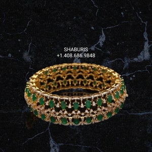 May include: A gold bangle bracelet with a delicate design featuring emerald green gemstones and smaller clear gemstones. The bracelet is on a black background.