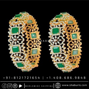 May include: Two gold bangles featuring intricate floral designs and emerald green gemstones. The bangles are set with small, clear diamonds.