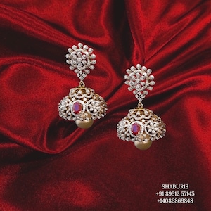 May include: A pair of gold earrings with a red stone and diamond accents. The earrings are in the shape of a jhumka, a traditional Indian style of earring.
