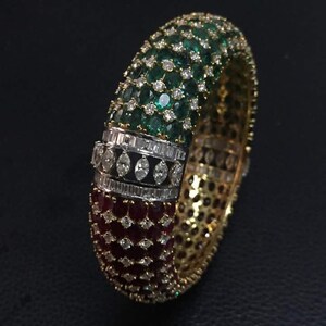 May include: A gold bangle bracelet featuring a band of emerald green, white, and ruby red gemstones. The bracelet is set with small round stones and a row of baguette-shaped stones.