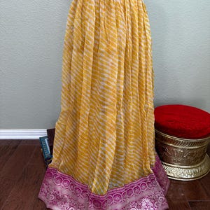 May include: A yellow and white patterned skirt with a pink and silver trim. The skirt is pleated and has a yellow waistband.