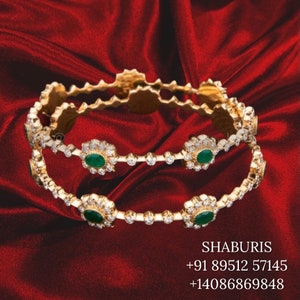 May include: Two gold bangles with emerald and diamond detailing are displayed on a red satin background. The bangles have a floral design with oval green emeralds and small diamonds. The text "SHABURIS" is visible.