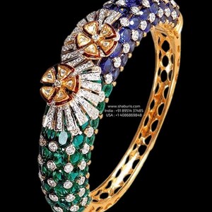May include: A gold bangle bracelet with a decorative design featuring three flower-shaped clusters of gemstones. The flowers are set with diamonds, sapphires, and emeralds. The bracelet is also set with smaller diamonds.