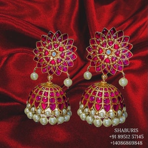 May include: A pair of gold earrings with pink and white stones. The earrings are in the shape of a flower with a round, dangling bottom. The earrings are on a red background.