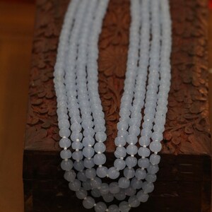 May include: A light blue beaded necklace with multiple strands of beads. The necklace is displayed on a wooden box with intricate carvings.