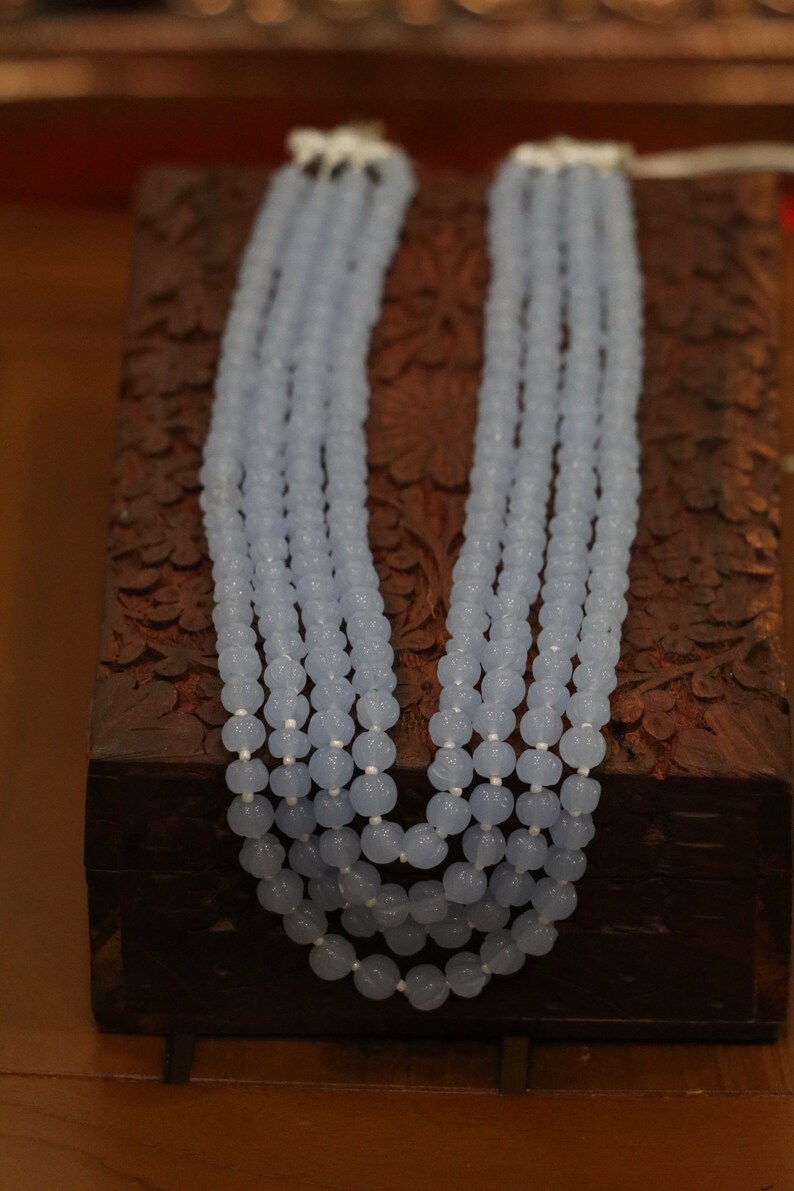 May include: A light blue beaded necklace with multiple strands of beads. The necklace is displayed on a wooden box with intricate carvings.