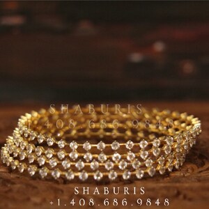 May include: Four gold bangles with clear stones set in a row around the bangle. The bangles are stacked on top of each other. The text "SHABURIS +1.408.686.9848" is visible below the bangles.