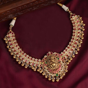 Pode incluir: Um colar de ouro com um pingente que apresenta uma divindade hindu, rodeado por pedras preciosas rosa e brancas. O colar está sobre um fundo bordô.