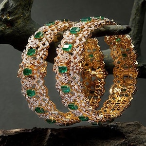 May include: Two gold bangles featuring intricate diamond and emerald designs. The bangles are set with emerald green stones and sparkling diamonds.
