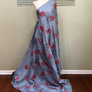 May include: A draped, light blue fabric with embroidered floral patterns in pink, purple, and green. The fabric is displayed on a mannequin, showcasing its texture and design. The floral pattern is evenly distributed across the fabric.