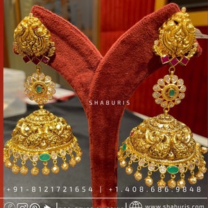 May include: A pair of gold earrings with intricate designs, featuring a peacock motif, green and red gemstones, and a dangling bell-shaped charm. The earrings are displayed on a red velvet background.