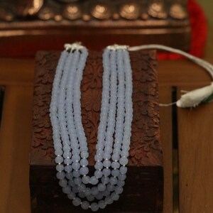 May include: A light blue beaded necklace with five strands of beads. The necklace is displayed on a wooden box with intricate carvings.