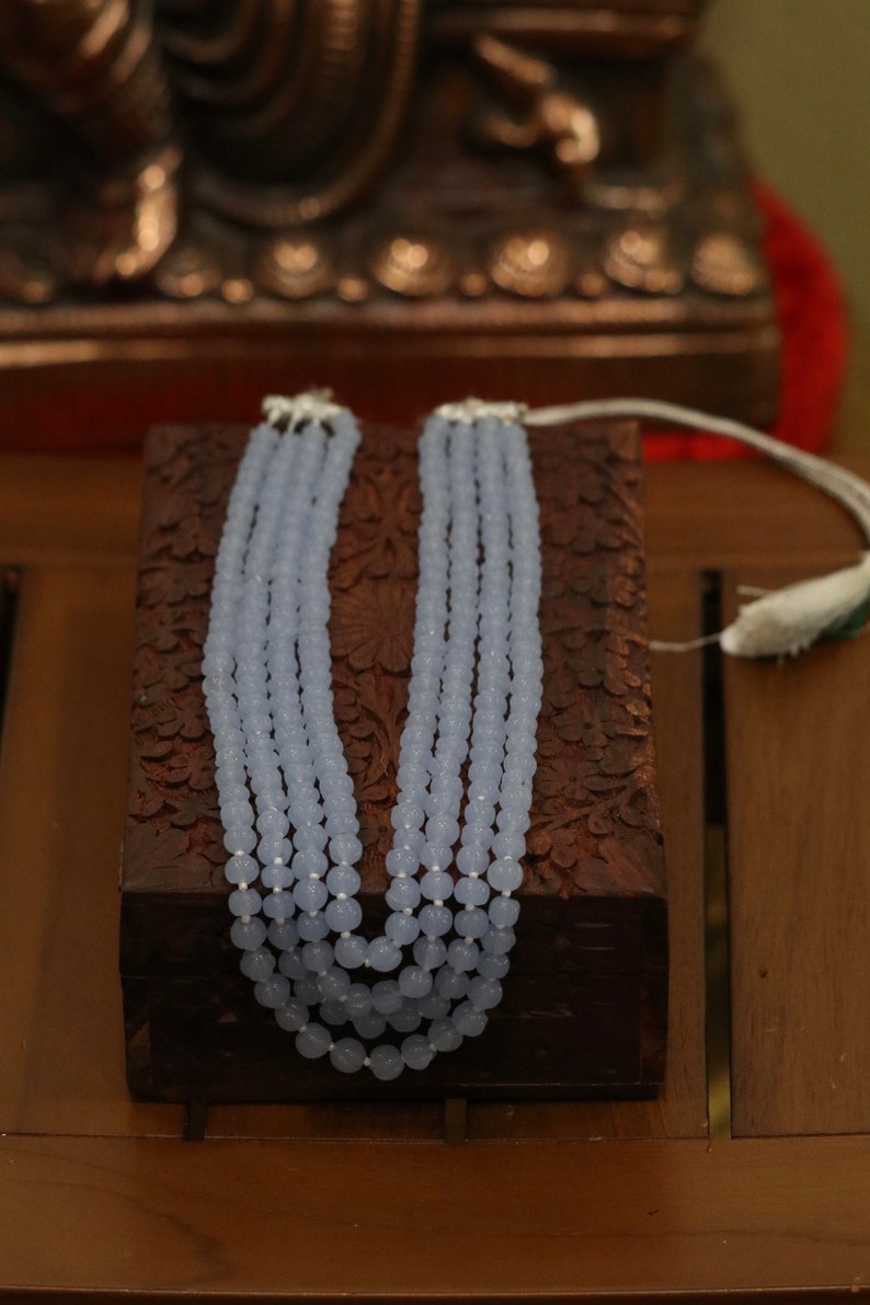 May include: A light blue beaded necklace with five strands of beads. The necklace is displayed on a wooden box with intricate carvings.