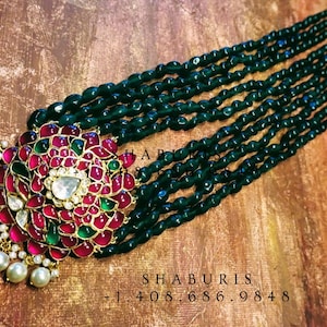 Puede incluir: Un collar de múltiples hileras con cuentas de color verde oscuro y un gran colgante adornado. El colgante está hecho de plata y presenta un diseño floral con gemas rojas y verdes. El collar está adornado con perlas blancas.