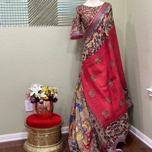 May include: A red and cream linen saree with a colorful floral and paisley print. The saree is draped on a mannequin and has a matching blouse with a short sleeve.
