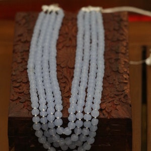 May include: A multi-strand necklace made of light blue beads. The necklace is displayed on a wooden box with intricate carvings.