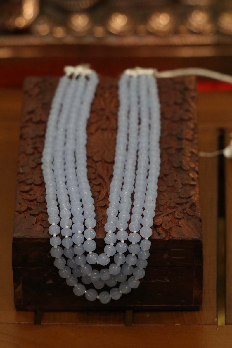 May include: A multi-strand necklace made of light blue beads. The necklace is displayed on a wooden box with intricate carvings.