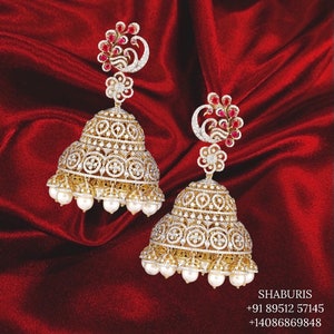 May include: A pair of gold earrings featuring a peacock design and white pearls. The earrings are set with sparkling diamonds and red gemstones.