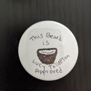 May include: White round button with the text "This Beard is Lucy Tollefson Approved!" and a drawing of a beard made of a coconut.