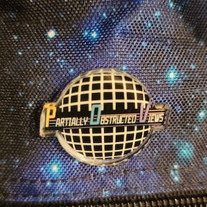 May include: A black and white grid pattern on a round, clear button with the text "Partially Obstructed Views" in blue, yellow, and purple. The button is attached to a blue and black galaxy-patterned fabric.