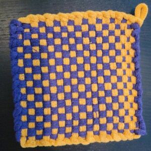 Puede incluir: Un posavasos tejido amarillo y morado con un patrón de cuadros y un lazo para colgar.