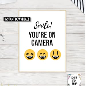 Peut inclure: Affiche imprimable blanche avec du texte noir qui dit "Sourire ! Vous êtes à la caméra" et trois smileys jaunes. L'affiche a un cadre en bois brun.