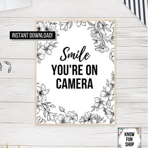 May include: A framed print with the words "Smile YOU'RE ON CAMERA" in black, surrounded by floral line art. The phrase "Instant Download!" is in a black banner. A small graphic in the bottom right corner says "KNOW FUN SHOP DIGITAL FILE".