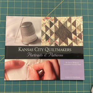 Puede incluir: Un libro titulado "Kansas City Quiltmakers Portraits & Patterns" con imágenes de herramientas de acolchado y patrones de edredones. La portada presenta un dedal, una aguja y un diseño de edredón con triángulos marrones, beige y verdes.