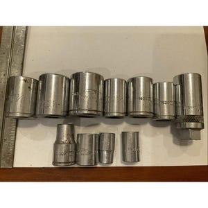 Puede incluir: Una colección de once accesorios de llave de vaso de metal plateado de varios tamaños, dispuestos en dos filas junto a una regla de metal que muestra medidas métricas. Cada vaso está grabado con números de modelo y marcas específicas.
