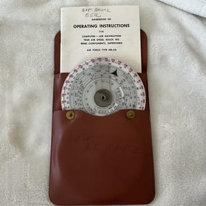May include: A brown leather case holds a white booklet and a circular slide rule. The booklet reads "Handbook of Operating Instructions" for air navigation, true air speed, and wind components. The slide rule has various scales and markings.
