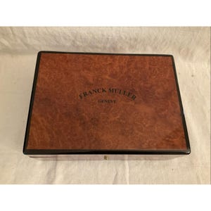 Może przedstawiać: Drewniane pudełko z czarną obwódką i brązowym wzorem słojów drewna. Pudełko ma mosiężny zatrzask i napis "FRANCK MULLER GENEVE" nadrukowany na górze.