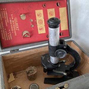 May include: A vintage Gilbert 5-16 microscope in a grey case with a red interior. The microscope is black and silver, with a focus knob and a rotating turret. The case also contains slides, a cork, and other accessories. The text on the case reads "GILBERT" and "MICROSCOPE".