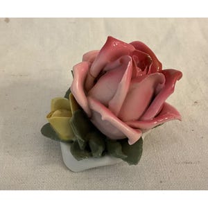 May include: A porcelain rose figurine with pink and white petals, yellow bud, and green leaves. The rose is sitting on a white base.