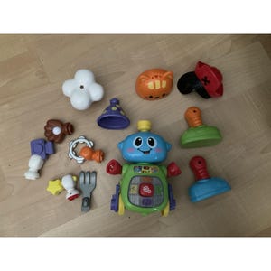 May include: A central blue and green interactive robot toy surrounded by various plastic accessories including a white cloud, orange cat head, red pirate hat, purple cone, green and blue bases, and small tools like a fork and camera.