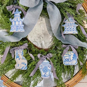 May include: A blue and white porcelain-inspired Christmas wreath with a blue and white gingham ribbon. The wreath is decorated with five ornaments: a pagoda, a vase, a dog, and two more pagodas. The wreath is hanging on a gold mirror with a decorative frame. The text "Photo & Styling by @homefernhill" is at the bottom of the image.