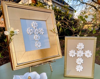May include: Two framed cross-shaped wall decor pieces with white ceramic flowers. The frames are gold with a white background. The flowers are arranged in a cross shape.