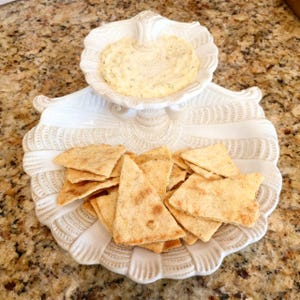 May include: A white, shell-shaped serving dish with a smaller dish on top, filled with a creamy dip. The lower dish holds several triangular crackers. The dish is on a speckled, brown surface.