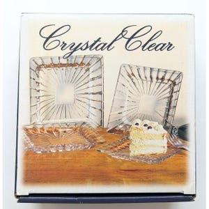 May include: A product box labeled Crystal Clear featuring four square glass plates with a sunburst pattern. One plate holds a slice of layered cake with cream frosting and berries. The box displays the plates on a wooden surface background.