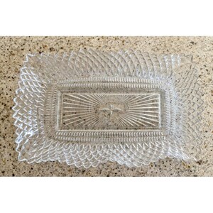 May include: A rectangular clear glass serving tray featuring a textured diamond pattern along the outer border and a central sunburst design radiating from a small four-pointed star shape in the middle of the flat base.