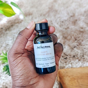 Puede incluir: Un peque&ntilde;o frasco de vidrio marr&oacute;n oscuro con una tapa negra, etiquetado como "The Potter's Mixtress Kash's Custom Serum" y que enumera ingredientes como aceite de aguacate, aceite de nuez de macadamia y aceite esencial de lavanda. El frasco est&aacute; etiquetado como "30ML/1OZ" y es org&aacute;nico, sin refinar, vegano y sin qu&iacute;micos.