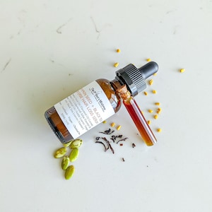 May include: A brown glass bottle with a dropper top containing Pumpkin Seed + Black Tea Reverse Hair Loss Serum. The bottle is labeled with the product name and ingredients. The bottle is sitting on a white surface with some dried tea leaves and pumpkin seeds.