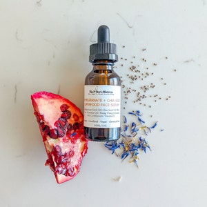 May include: A brown glass bottle of face serum with a dropper, labeled "Pomegranate + Chia Seed Superfood Face Serum." A pomegranate slice, chia seeds, and blue cornflowers are arranged around the bottle. The bottle contains 1 fl oz of serum.