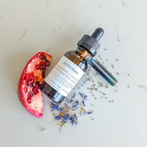 May include: A brown glass bottle of "Pomegranate + Chia Seed Superfood Face Serum" with a dropper, a pomegranate half, chia seeds, and blue cornflowers. The bottle is labeled with product information. The serum is 1 fl oz.