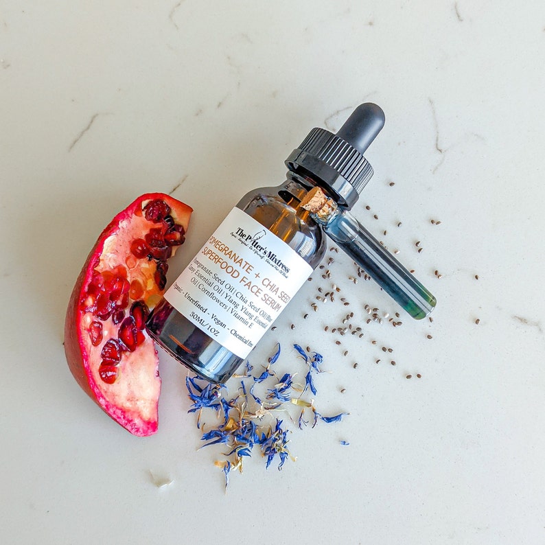 May include: A brown glass bottle of "Pomegranate + Chia Seed Superfood Face Serum" with a dropper, a pomegranate half, chia seeds, and blue cornflowers. The bottle is labeled with product information. The serum is 1 fl oz.