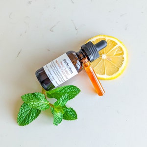 May include: A brown glass bottle with a dropper top containing The Pfter's Mistress Triple Threat Radiance Face Serum. The bottle is labeled with the product name and ingredients, including Rosehip Seed Oil, Sea Buckthorn Oil, Japanese Green Tea, and Vitamin E. The bottle is sitting on a white surface with a lemon slice and a sprig of green mint.