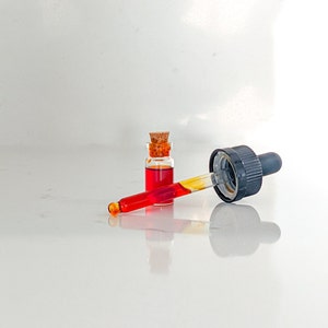 May include: A glass bottle with a cork stopper filled with a red liquid and a glass dropper with a black rubber tip.