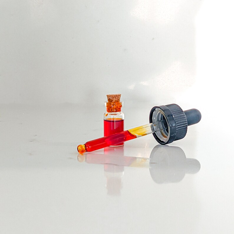 May include: A glass bottle with a cork stopper filled with a red liquid and a glass dropper with a black rubber tip.