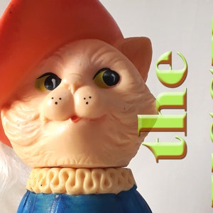 Puede incluir: Un gato de juguete de plástico vintage que lleva un sombrero rojo y un atuendo azul y blanco. El gato tiene un bigote blanco y una barba blanca. El texto "the USSR" está escrito en letras verdes sobre un fondo blanco.