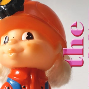 May include: A plastic toy figurine of a child wearing an orange hard hat with a black headlamp, a red and blue jumpsuit, and blonde hair. The text "the USSR" is written in pink letters in the background.